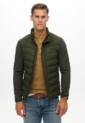 Mężczyzna w oliwkowej kamizelce puchowej na khakowym długim rękawie, niebieskich dżinsach, brązowym pasku i zegarku na nadgarstku, stojący na tle białego tła.