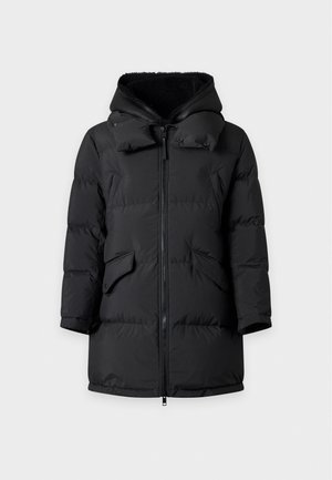 Homme portant une doudoune noire à capuche, un polo beige, un pantalon vert foncé et des baskets blanches, debout devant un fond clair uni.