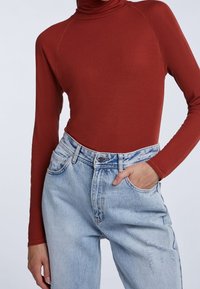 Maglione a collo alto in ruggine con maniche lunghe abbinato a jeans a vita alta, chiari e strappati, dotati di cinque tasche e chiusura con bottone.