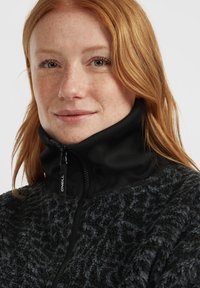 Close-up van een roodharige vrouw met sproeten, gekleed in een zwart fleece jack dat tot aan de hals is dichtgeritst, tegen een effen achtergrond.