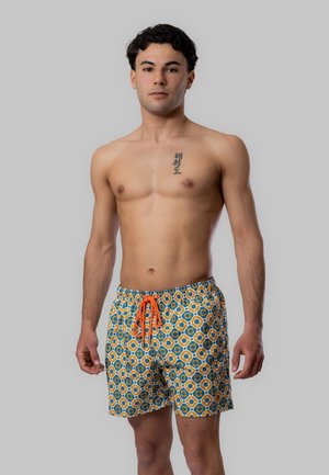 Giovane uomo con capelli neri ricci, senza maglietta, indossa pantaloncini da bagno blu, gialli e arancioni con un laccio arancione, in piedi davanti a uno sfondo grigio.