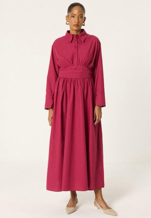 Femme debout dans une robe magenta à manches longues avec une large ceinture, un haut boutonné à col et des talons bas beige pointus.