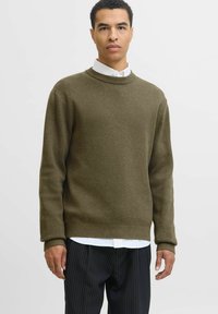 Vīrietis, kurš valkā olīvzaļo džemperi pār baltu krekla un melnajām svītrainajām biksēm, stāvot pret vienkāršu gaišu fona.