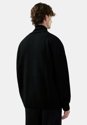 Uomo con capelli ricci che indossa un maglione nero oversize e pantaloni neri con texture, di spalle su uno sfondo chiaro e uniforme.