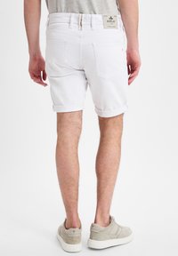 Vita denimshorts med uppvikta fållar, två bakfickor och en synlig varumärkesetikett ovanför midjan. Bärs med ljusgrå sneakers.