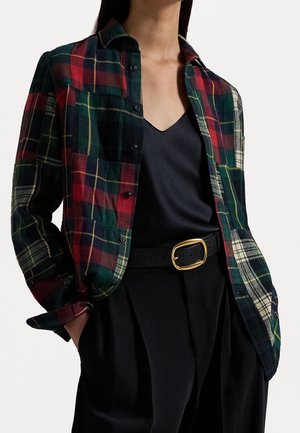 Femme portant une chemise à carreaux rouge, verte et jaune, déboutonnée, par-dessus un haut noir à col en V, un pantalon noir et une ceinture noire avec une boucle dorée, les mains dans les poches.