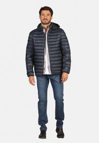 Doudoune marine avec fermeture éclair et capuche, présentant un design matelassé. Associée à une chemise blanche et un jean bleu, ainsi qu'à des bottes noires.