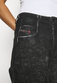 Jeans de mezclilla negra con una textura desgastada, un patrón de salpicaduras blancas y una etiqueta de marca visible en el bolsillo frontal. Corte estándar.
