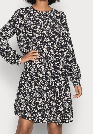 Robe longue à manches longues noire avec imprimé floral blanc et beige, encolure ronde et jupe froncée, portée par une personne avec une main sur la hanche.