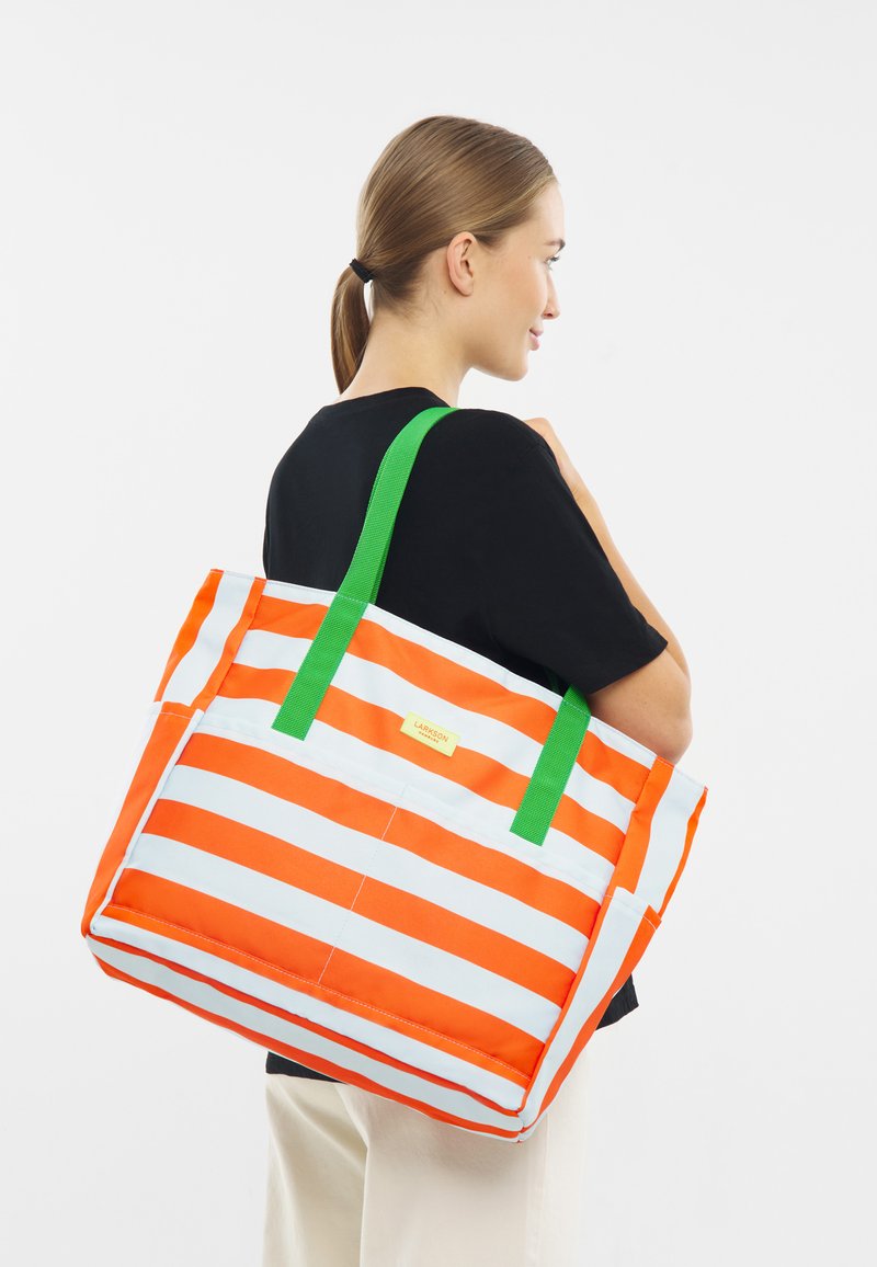 Donna in camicia nera che porta una grande borsa a tracolla arancione e bianca a strisce con manici verdi, su uno sfondo bianco.