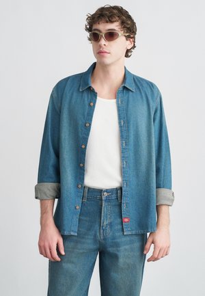 Chemise en jean boutonnée de couleur bleu clair avec manches retroussées, associée à un jean taille haute assorti. Le mannequin porte un débardeur blanc et des lunettes de soleil.