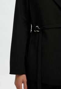 Veste noire structurée au design enroulé, dotée d'une ceinture nouée et d'accents en métal argenté. Tissu lisse et manches longues sont visibles.
