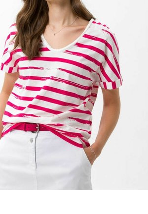 Woman wearing a white and pink striped short-sleeve V-neck shirt tucked into a white skirt with a pink belt, hand in pocket.