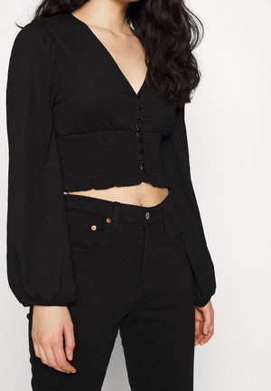 Kobieta ubrana w czarny, długi crop top z guzikami z przodu oraz czarne jeansy z wysokim stanem na białym tle.