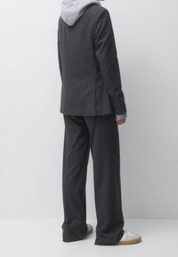 Traje gris a rayas con un ajuste ligeramente holgado, combinado con una sudadera gris con capucha. Presenta una abertura trasera y pantalones de pierna ancha. Zapatillas blancas completan el look.