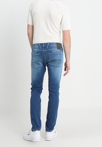 Man som bär urtvättade blå jeans, en vit kortärmad tröja i stickad stil och vita sneakers, står vänd bort från kameran mot en neutral bakgrund.