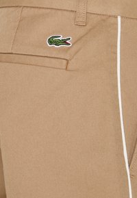 Beżowe bawełniane spodnie o teksturowanym wykończeniu, z białym paskiem bocznym i zielonym logo krokodyla na tylnej kieszeni.