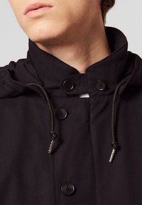 Czarna kurtka z wysokim kołnierzem, z dwoma czarnymi guzikami, sznurkiem z srebrnymi akcentami oraz fakturowanym materiałem.