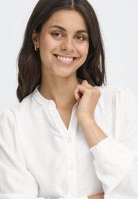 Camicia bianca a maniche lunghe con colletto alla cinese, pattina e maniche elasticizzate. Texture liscia, design minimalista.