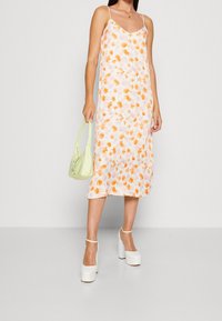 Žena ve bílé midi slip dress s oranžovým květinovým vzorem, držící světle zelenou kabelku a obutá v bílých platformových podpatcích.