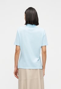 Γυναίκα με σκούρα μαλλιά μέχρι τους ώμους, που φοράει ανοιχτό μπλε κοντομάνικο τοπ και μπεζ φούστα, πλάτη σε μονόχρωμο φόντο.