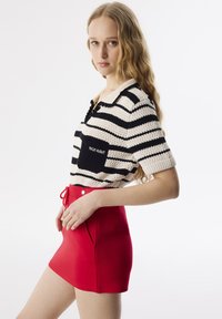 Haut en tricot rayé crème et noir avec col, comportant une poche poitrine. Associé à une mini-jupe rouge unie avec poches latérales et cordon de serrage.
