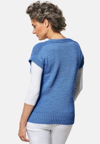 Blauwe gebreide trui met opgerolde korte mouwen, textuurpatroon en geribbelde zoom, gecombineerd met een witte langemouwtop eronder.