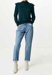 Kvinna i mörkgrön långärmad tröja, ljusblå uppvikt jeans och beige klackade ankelskor, stående med händerna i fickorna på vit bakgrund.