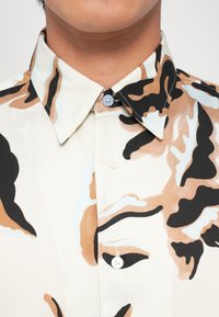 Košile na knoflíky s krémovým základem, zdobená hnědým a černým květinovým vzorem. Límeček je špičatý, s kontrastními modrými a bílými akcenty.