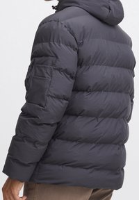 Chaqueta acolchada en gris oscuro, con capucha, diseño acolchado horizontal y un bolsillo con cremallera en la manga izquierda. Textura suave y acolchada.