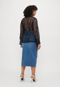 Top in pizzo blu navy con maniche lunghe e dettagli con fiocchi ai lati, abbinato a una gonna midi in denim. I materiali presentano intricate fantasie in pizzo e denim liscio.