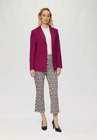 Fuchsias Blazer, tailliert mit Revers; darunter ein weißer Pullover. Gemusterte, verkürzte Hose in Rot, Schwarz und Weiß. Schwarze hohe Absätze.