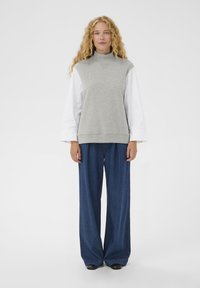 Pull-over sans manches gris à col montant, superposé sur une chemise blanche à manches longues. Associé à un pantalon large en denim bleu et à des chaussures noires.