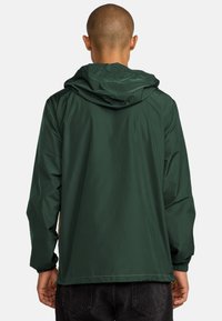 Giacca leggera verde scuro con cappuccio, dotata di polsini elastici e una superficie liscia e lucida. Design semplice e minimalista senza hardware visibili.