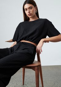 Černý texturovaný dvoudílný outfit skládající se z cropped topu s krátkými rukávy a volných kalhot. Model sedí na dřevěné židli s neutrálním pozadím.