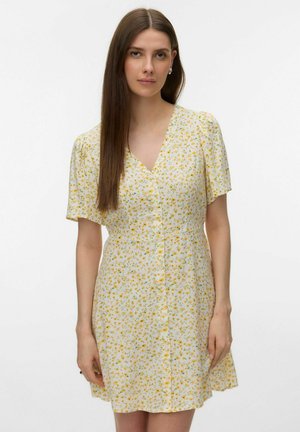 Mujer con cabello largo, liso y castaño, que lleva un vestido blanco de manga corta con un pequeño estampado floral amarillo y verde, sobre un fondo liso.