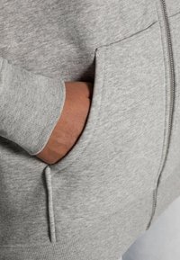 Sweat à capuche gris en tissu doux et texturé avec une fermeture éclair, doté d'une poche latérale et de poignets côtelés. Gros plan sur une main dans la poche.