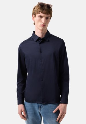 Junger Mann mit kurzem braunem Haar, der eine Sonnenbrille auf dem Kopf trägt, ein marineblaues Hemd mit Knöpfen und hellblaue Jeans vor einem einfarbigen Hintergrund.