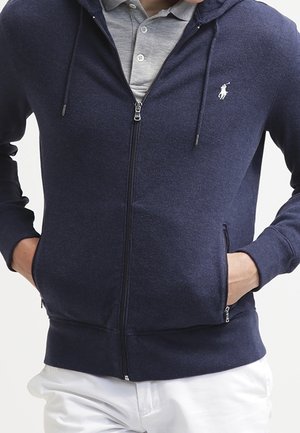 Mężczyzna ubrany w granatową bluzę z kapturem na zamek, z białym logo na piersi, pod spód szara koszula z kołnierzykiem oraz białe spodnie, z rękami w kieszeniach.