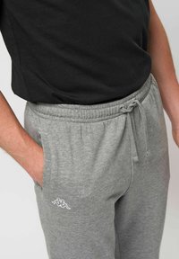 Person iført grå joggingbukser med snøre og sidelommer, hånd i lommen, og en sort skjorte stikket ind.