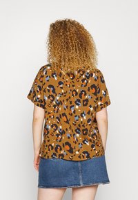 Haut à manches courtes, imprimé léopard marron avec des accents orange, bleu et blanc, porté avec une jupe en jean affichant des coutures visibles.