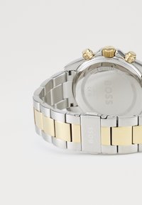 Silver and gold-tone metal watch with a linked bracelet, three chronograph pushers, and a clear back showing "BOSS" branding.