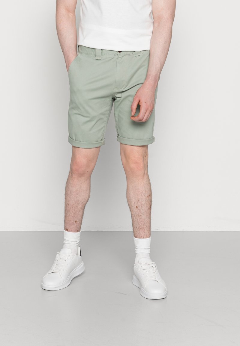 Shorts en coton vert clair, avec des ourlets roulés, poches latérales, associés à des sneakers blanches et des chaussettes, portés avec un t-shirt blanc uni.