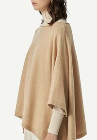 Frau trägt beigen Wollponcho über cremefarbenem Rollkragenpullover, steht im Seitenprofil vor einem schlichten Hintergrund.