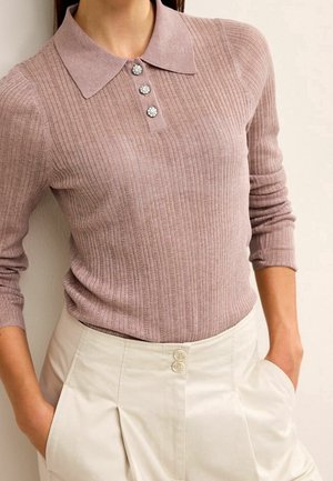 Femme portant un pull polo côtelé mauve à manches longues avec trois boutons décoratifs et un pantalon crème taille haute avec poches.