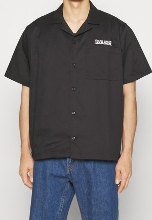 Chemise noire à manches courtes en tissu lisse, dotée d'une poche avant et d'un logo sur la poitrine gauche. Fermeture à boutons sur le devant.