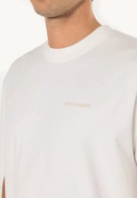 Camiseta blanca de algodón con cuello redondo. Presenta un sutil logo color beige "PESCADOR" en el pecho. Textura suave, corte relajado.