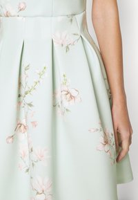 Robe vert clair avec imprimé floral rose, jupe plissée, et le bras d'une personne reposant à côté.