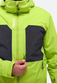 Veste imperméable verte vif avec un panneau à motif à carreaux noir. Comprend une fermeture éclair devant, une capuche ajustable et des poignets élastiques.