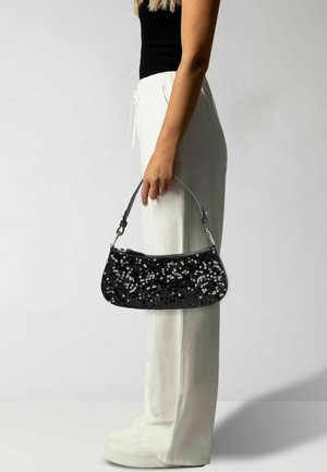 Sac à main noir en sequins avec une forme arrondie, fermeture éclair et poignée courte, tenu par une personne en pantalon blanc.